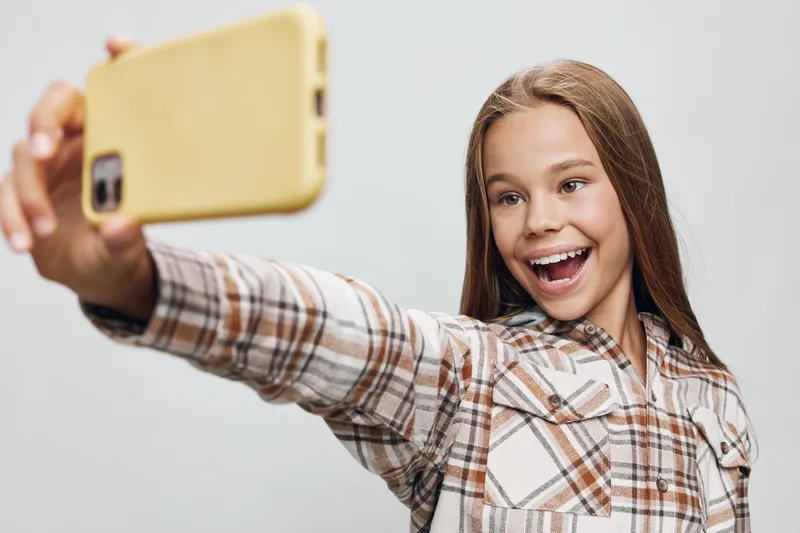 Girl taking selfie with smartphone smiling with excitement while wearing plaid shirt on neutral background. Portrait of happy teen holding phone for photo in casual style.