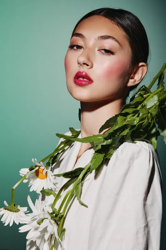Creative beauty portrait of a young woman with glossy red lips and rose-colored blush, holding white daisies over white clothes against a turquoise background. Fashion concept.