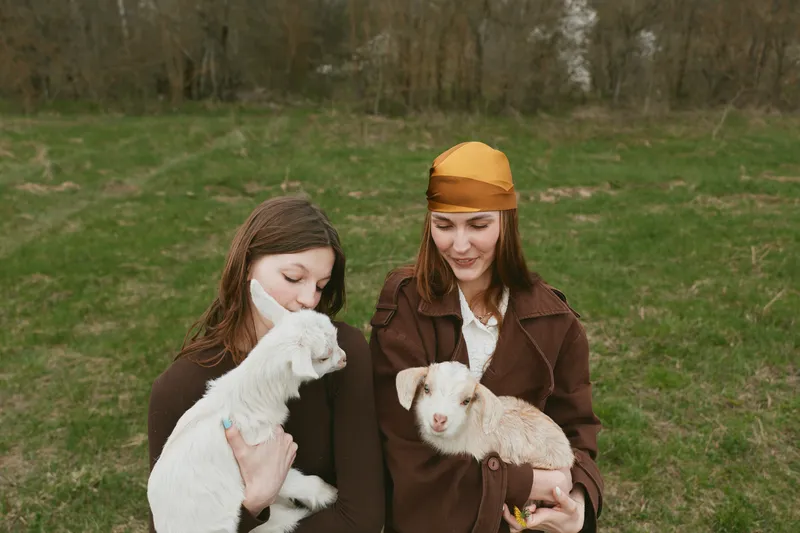 Goat kid held by two women outdoors, in a grassy field, highlighting farm lifestyle, joy and affection as they cradle friendly animals in a peaceful rural setting with soft mood.