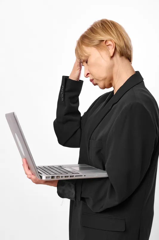 Stressed woman in dark office wear, holding a laptop, hand on head with headache, anxious expression, concept of burnout, stress, mental health, modern office technology