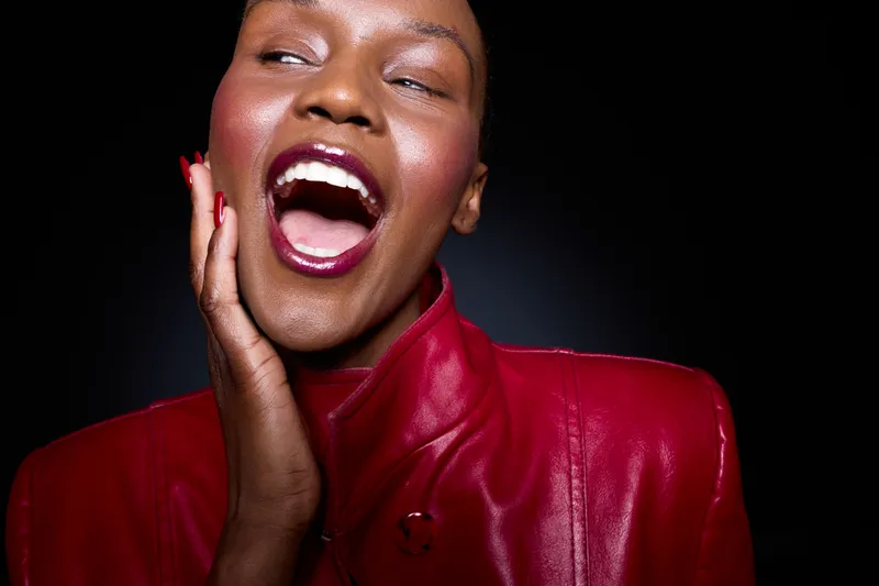 Joyful black woman with dark skin wearing a red leather jacket laughs with her mouth open and hand on cheek. Studio portrait on black background. Concept of happiness and fashion.