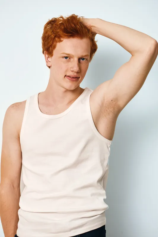 Young man with red hair wearing white tank top posing with arm raised showing underarm against light background. Casual portrait with relaxed expression and natural look.