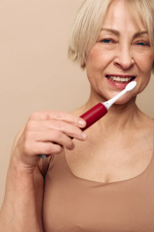 Smiling mature woman with short blonde hair holds red electric toothbrush near mouth, showing dental care routine. Clean beige background highlights healthy smile and oral hygiene concept.