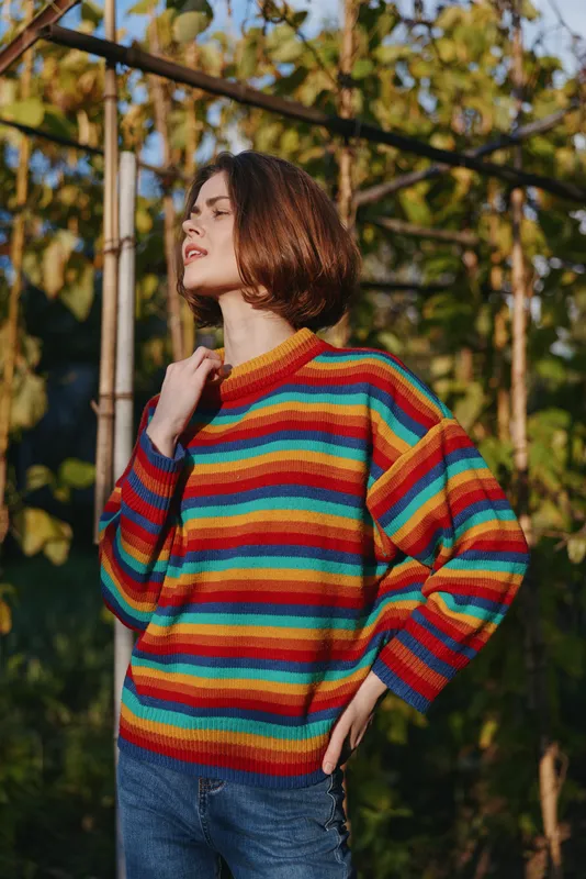 woman mid twenties with short hair wearing a rainbow striped sweater and jeans outdoors in a sunlit greenhouse, smiling confidently in a colorful casual outfit for fashion lifestyle portrait.