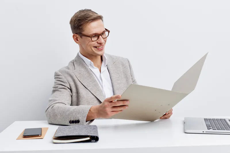 Smiling professional man in gray blazer reading documents at desk with laptop and notepad on white background. Business, office work, success and confident lifestyle concept.