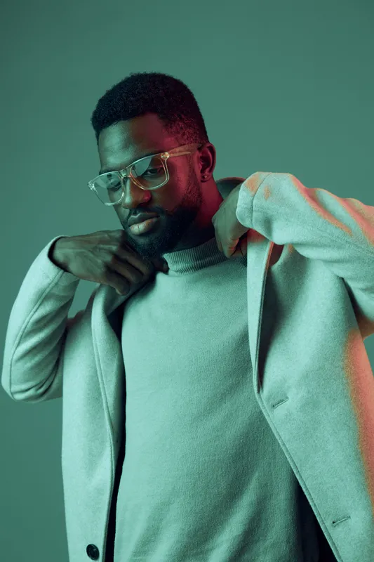 Stylish Black man adjusting his coat, wearing glasses, against a soft green background, showcasing a modern urban aesthetic and confident expression