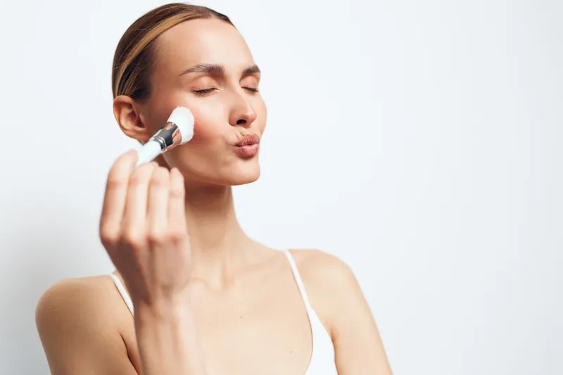 A young woman with clear skin applies makeup with a brush on her cheek, eyes closed and lips puckered. The bright neutral background emphasizes beauty and skincare concepts.