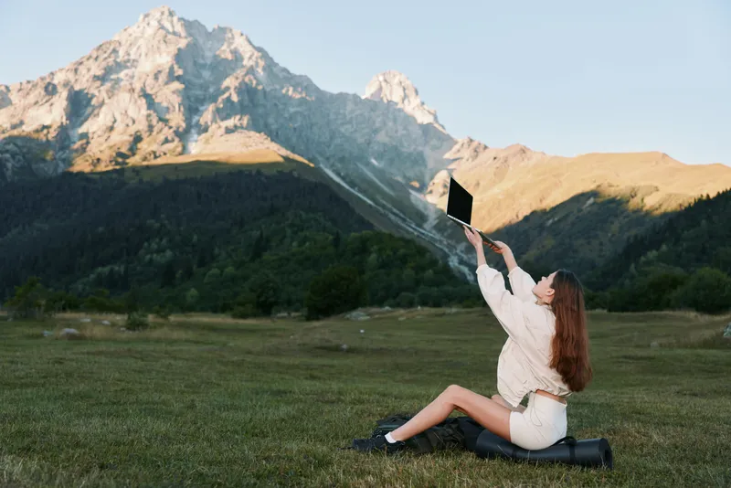 A cheerful woman sits on grass in a mountain landscape, holding a laptop above her head to capture a selfie. She smiles warmly, showcasing a bright, carefree outdoor work moment.