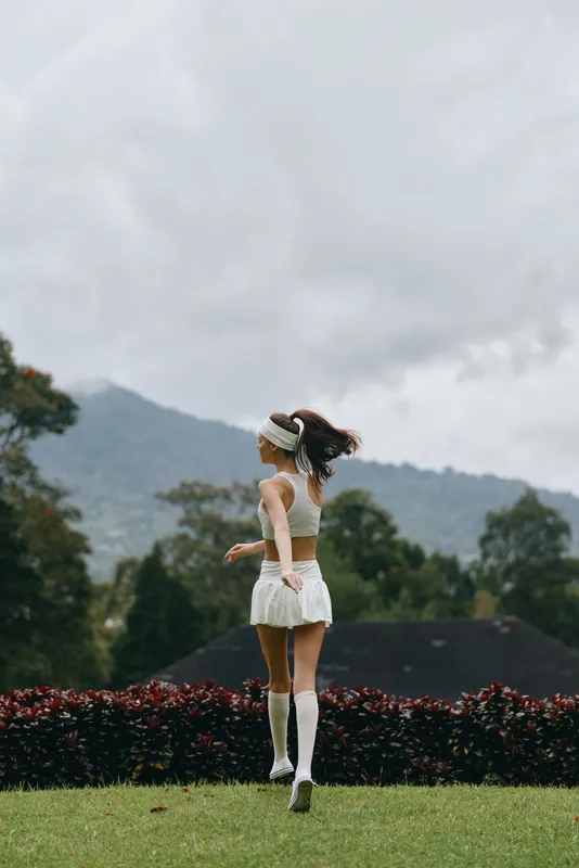 Young model in sports attire joyfully running outdoors, showcasing athleticism and vitality amidst a lush green landscape with a cloudy backdrop