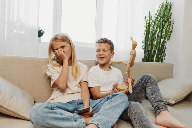 Two children sitting on a beige sofa in a bright living room, girl covering nose in disgust while boy holds wooden mannequin. Concept of childhood, sibling interaction, and playful emotions.