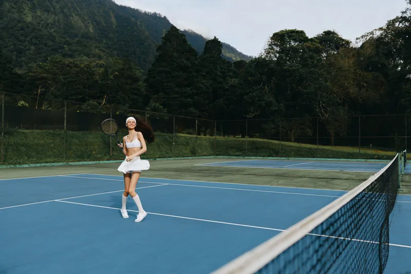 Young female tennis player in white sportswear expressing joy while playing on an outdoor court surrounded by green mountains and trees