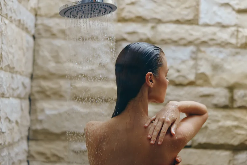 woman in shower with wet hair touching shoulder under water stream on stone wall background, natural light, relaxation and cleansing moment captured