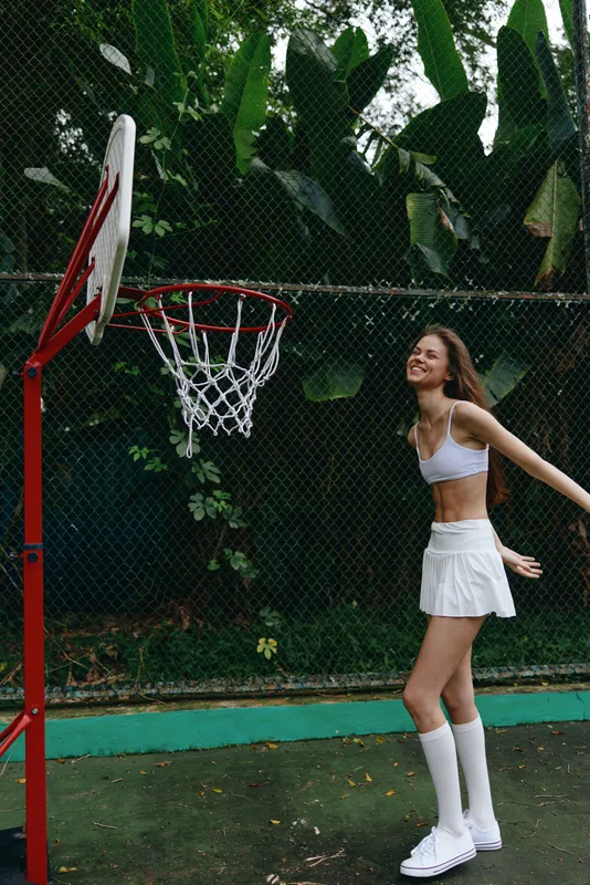 Young female athlete in a white sports outfit joyfully playing basketball outdoors, surrounded by lush greenery, conveying a sense of fun and vitality