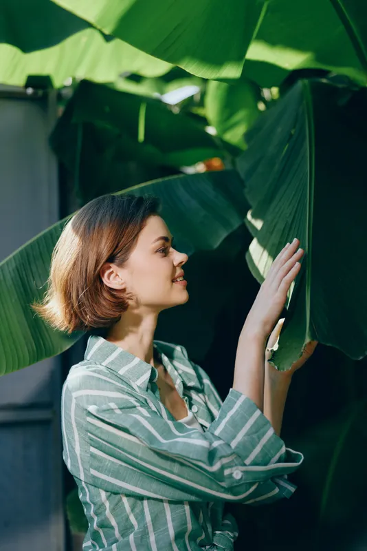A woman in a striped shirt gently touches large green leaves, showcasing calm beauty and natural grace. The quiet moment highlights connection with nature and a contemplative mood.