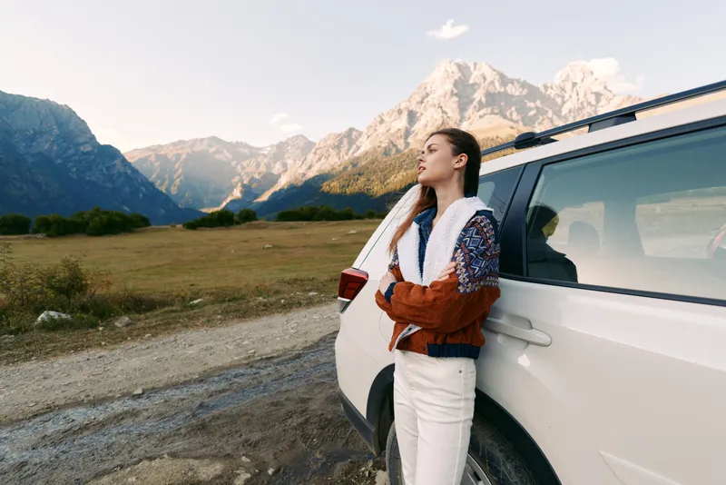 woman car mountains road travel nature landscape portrait. Young woman leans on white SUV at roadside, enjoying a scenic mountain view during a road trip, outdoor adventure and freedom.
