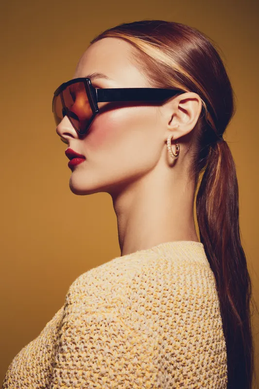 Profile of a stylish woman with long hair wearing large sunglasses and hoop earrings. She wears a textured sweater against a warm brown background. Fashion and beauty concept.