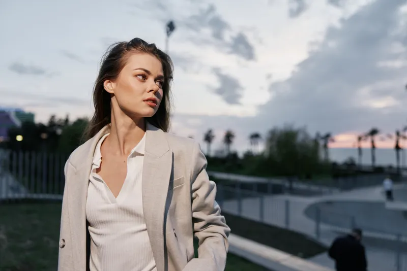 Woman in beige coat standing outdoors at sunset with thoughtful expression, casual style, urban background, natural light, autumn season, fashion concept
