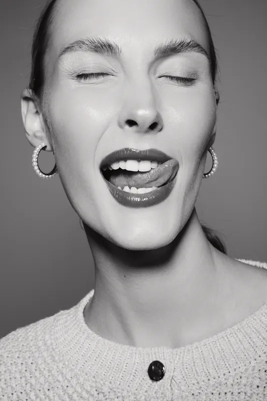 Portrait of a light skinned white woman with closed eyes and tongue out smiling, wearing earrings and a sweater, studio lighting on a neutral background.