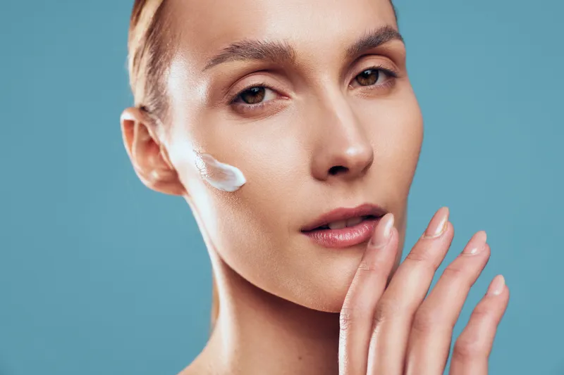Close-up of woman applying white face cream on cheek with fingers, clear skin and natural makeup on blue background. Concept of skincare, beauty, and facial care routine.