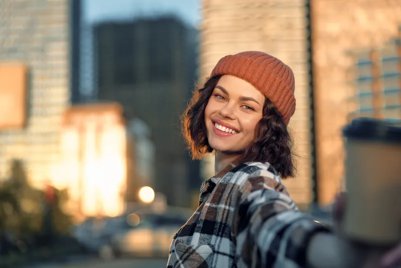Woman smile city selfie coffee candid lifestyle golden hour authenticity: warm urban portrait of a smiling woman in a cozy beanie at golden hour glow, mindful living and emotional storytelling.
