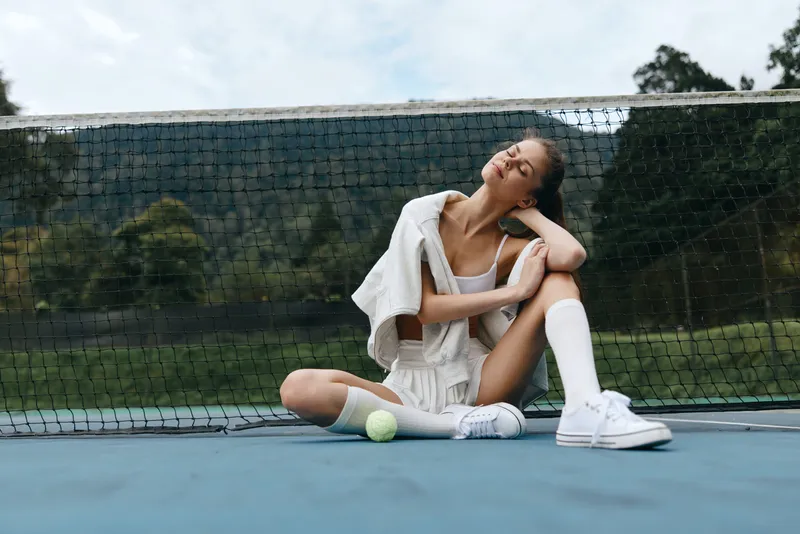 Young athletic woman in sportswear sitting on a tennis court, exuding confidence and joy, with a green nature backdrop and seasonal vibe of spring or summer