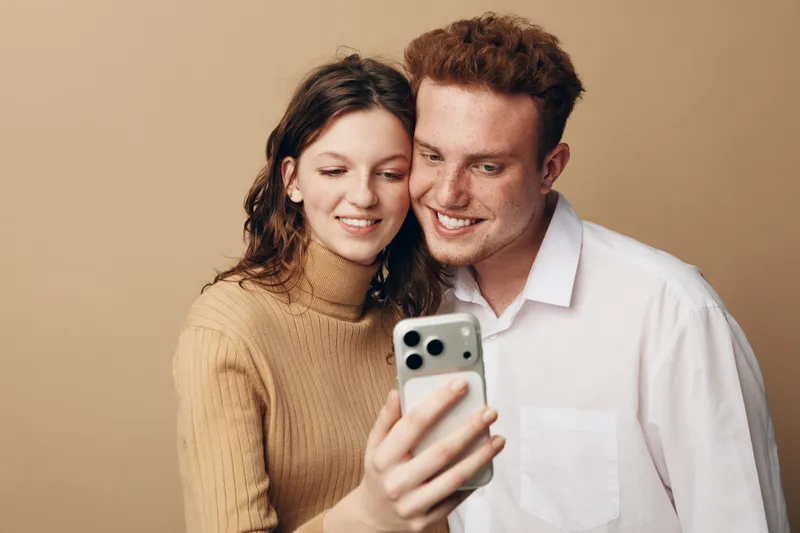 Smiling young couple taking selfie with smartphone, close together in casual clothes on beige background. Concept of friendship, technology, and happy moments shared.