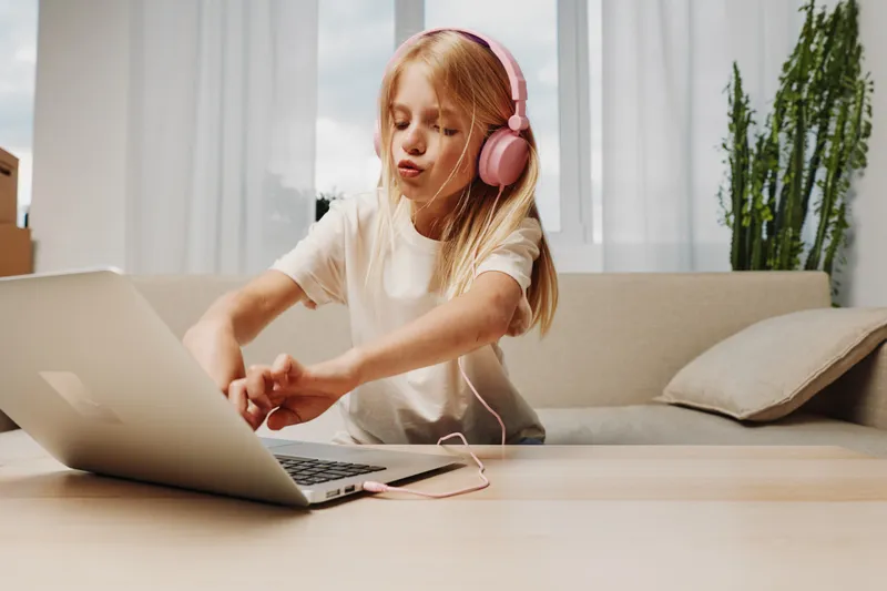 Blond girl with pink headphones enjoying music while using laptop at home. Casual outfit and bright modern living room background create cozy and relaxed atmosphere.
