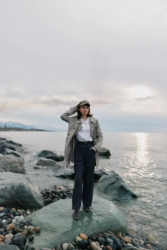 A young woman in a plaid coat and hat stands on rocks by the ocean during cloudy weather. The calm sea and distant horizon create a peaceful outdoor atmosphere.