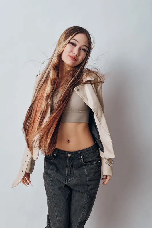 Young woman with long hair wearing beige jacket and cropped sweater posing confidently against white background with natural expression and casual style outfit.