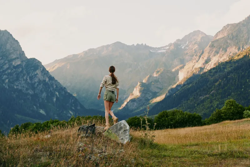 A woman in casual clothes stands on a rock in a grassy field looking at distant mountains under a clear sky, representing outdoor lifestyle and adventure.