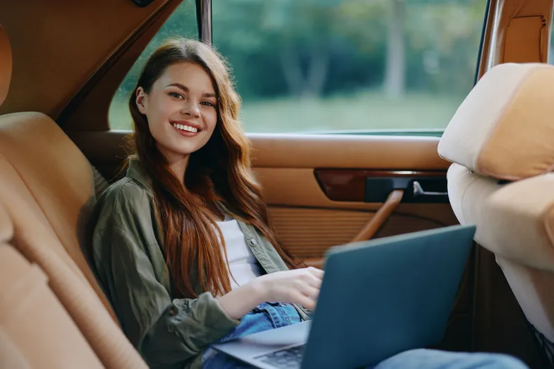 Young woman with long hair smiling while using a laptop in a car, showcasing a relaxed attitude and modern workspace concept.