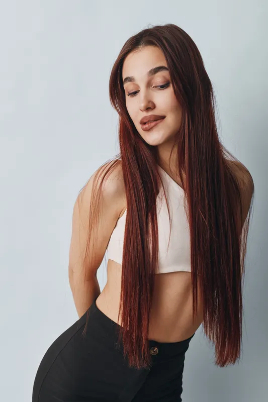 Young woman with long braided hair wearing a white crop top and black pants posing against a light background. Fashion model with natural makeup and confident expression.