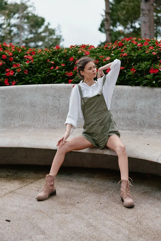 Stylish young woman in a green overall and white blouse sitting on a bench with red flowers in the background, embodying spring fashion and casual elegance