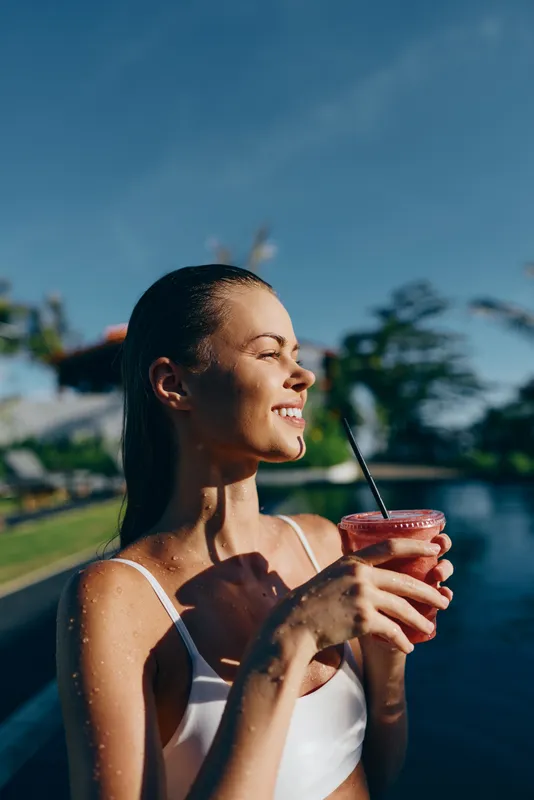 Young woman in white swimsuit enjoys fresh smoothie by the pool on sunny day outdoors smiling with eyes closed feeling relaxed and happy summer lifestyle concept