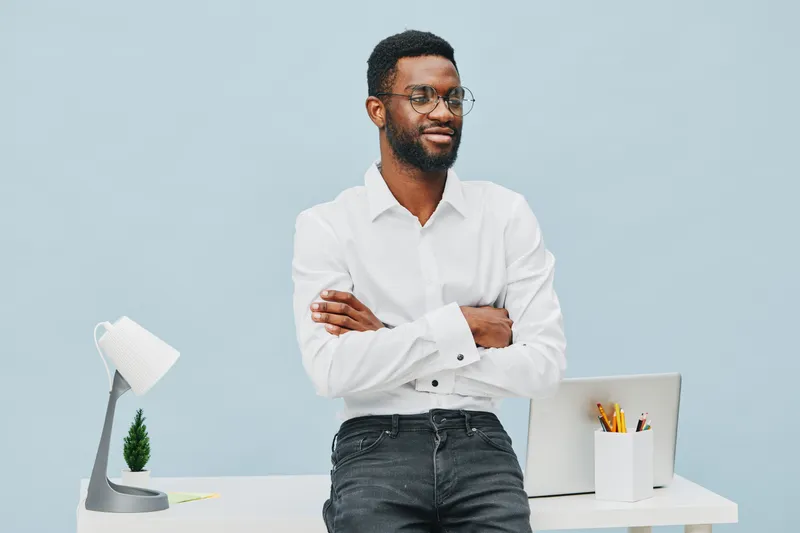 Confident young African-American man with beard and glasses in white shirt standing with arms crossed at modern desk with lamp and stationery on light blue background. People lifestyle concept