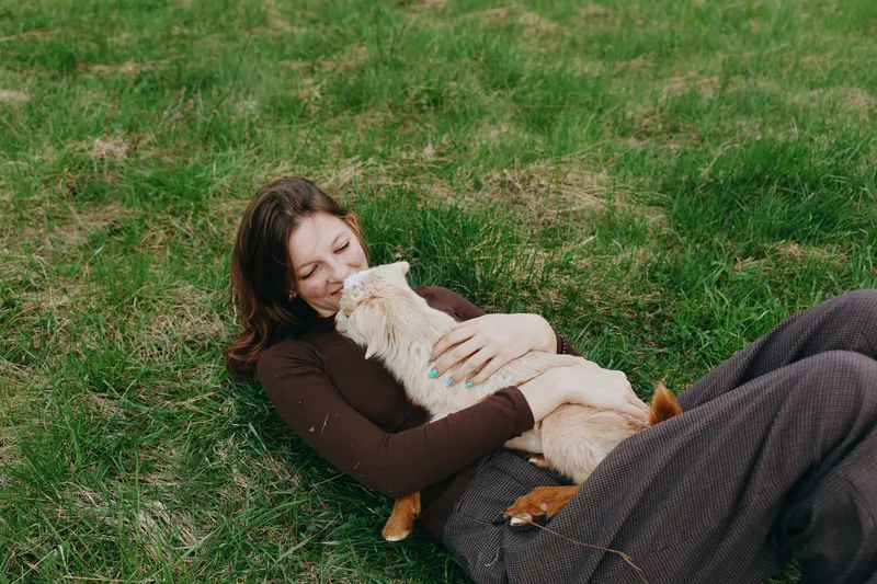 Lifestyle moment of a woman outdoors, lying on grass and cuddling a goat kid, gentle smile, soft breeze, warm tones, natural setting, relaxed mood, affectionate connection with nature.