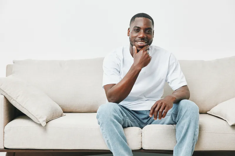 Happy young man smiling while sitting on a couch in casual wear, showcasing relaxation and confidence in a minimalist setting