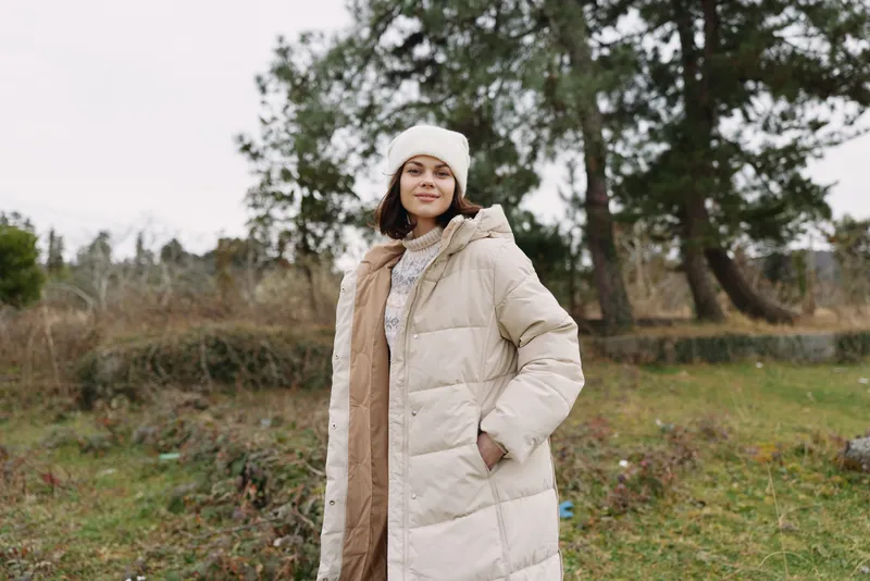 Winter girl wearing a beige puffer coat stands outdoors among trees, conveying a casual fashion moment in a tranquil natural landscape. Soft light and relaxed pose capture cozy style.