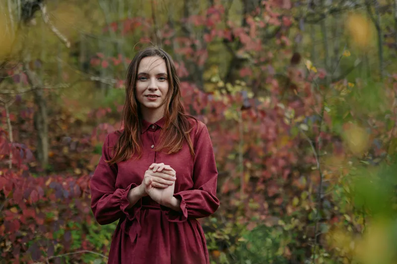 A candid portrait of a young woman in a deep burgundy dress standing in an autumn garden, radiating authenticity and quiet natural poise, warmth and confidence.