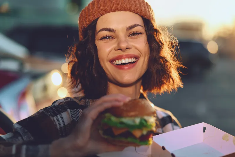 Woman smile burger outdoor beanie street food happy candid lifestyle portrait joyful young woman holding a burger at golden hour glow, authenticity and emotional storytelling.