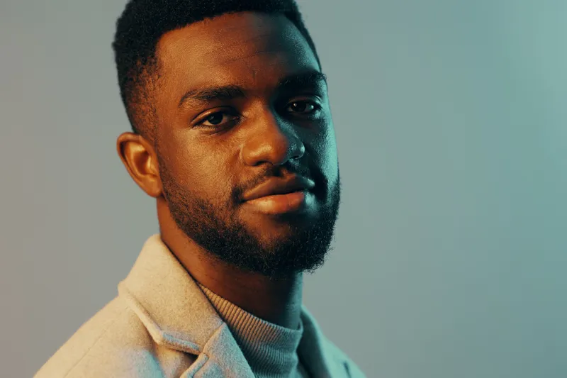 A confident young man in a stylish light colored coat against a soft blue and green background, showcasing modern fashion trends and a relaxed vibe