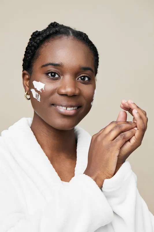 Smiling black woman applying skincare cream in a bright, minimalist setting, promoting self care and healthy skin