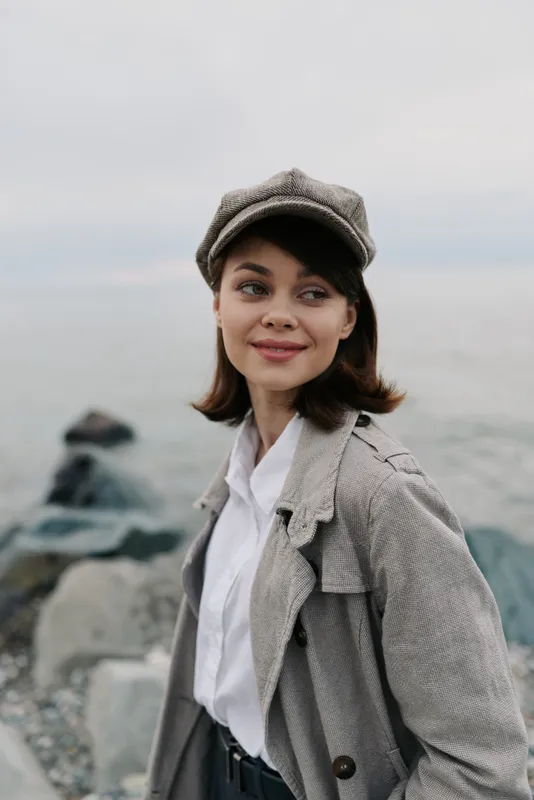 Young woman in vintage hat and textured coat smiles softly, standing near rocky seaside. Calm atmosphere with muted colors and natural light. Concept of casual style and outdoor portrait.