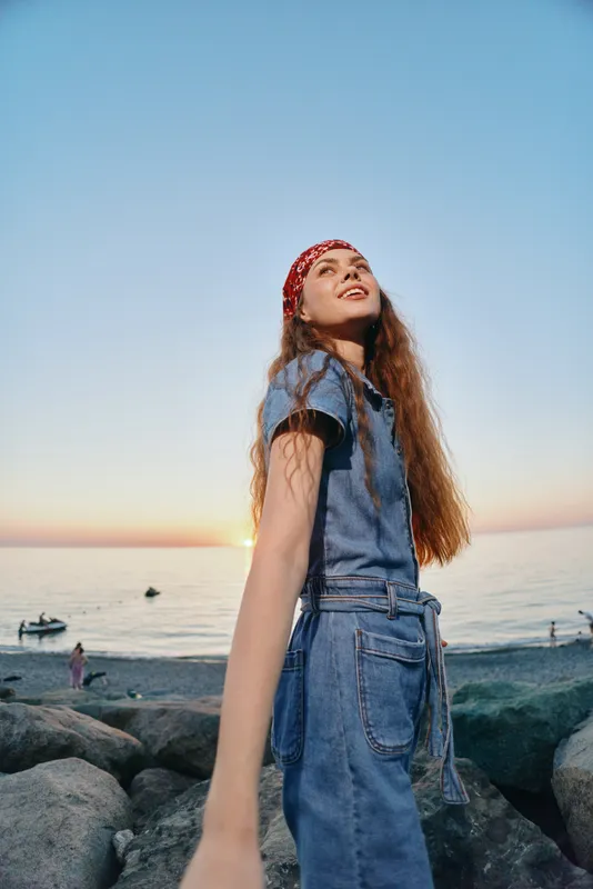 Lifestyle woman in modern boho-western denim street style smiling outdoors by the beach during sunset with film color effect and relaxed mood.