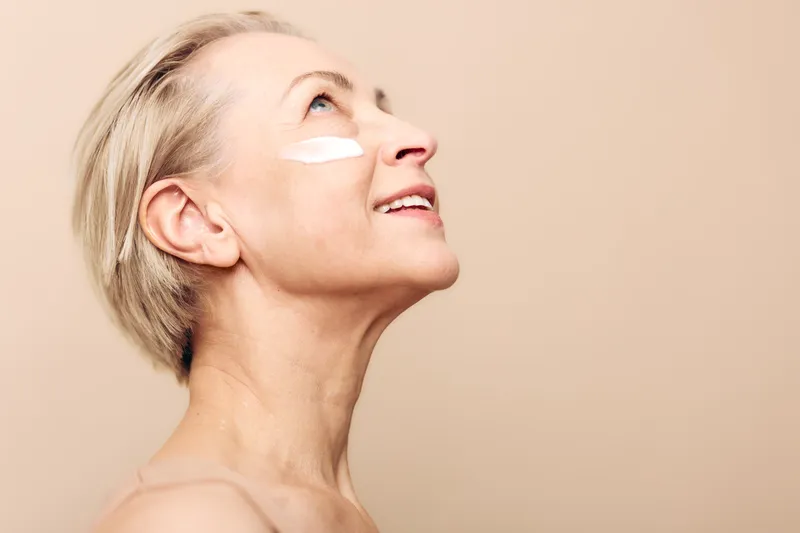 Mature woman with short blonde hair applies white cream on cheek, looking up against beige background. Concept of skincare, aging beauty, self-care, and healthy skin routine.