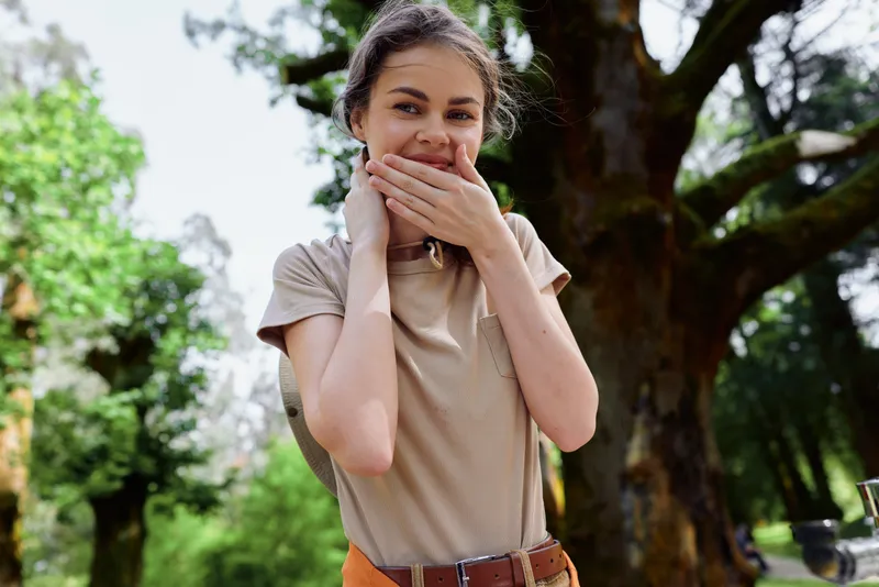 Woman girl portrait with surprise and smile in outdoor park, hands covering mouth in a candid emotional moment, young casual model showing joyful expression and relaxed natural background.
