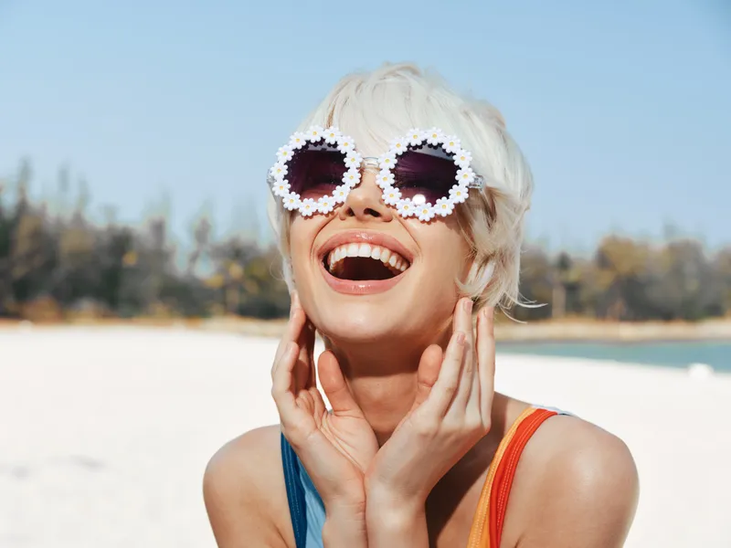 Bright joyful woman on a sunny beach wearing oversized flower sunglasses, white hair, and a colorful swimsuit, laughing with hands near her face. Her vibrant smile captures carefree summer energy.