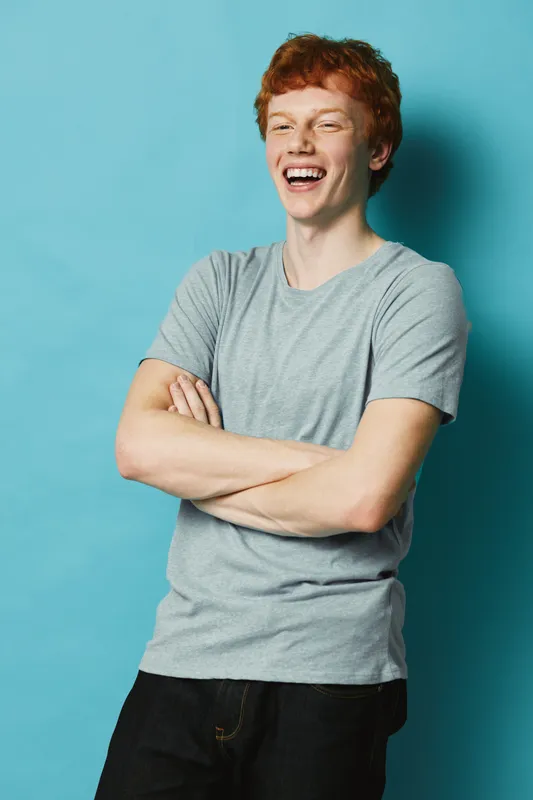 Young man with red hair smiling and laughing with arms crossed wearing casual t shirt and black pants standing against blue background in studio portrait.