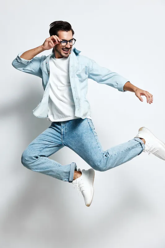 Full body happy cheerful young man jumping with excitement wear denim jacket and glasses isolated on plain light background. Joyful expression lifestyle concept.