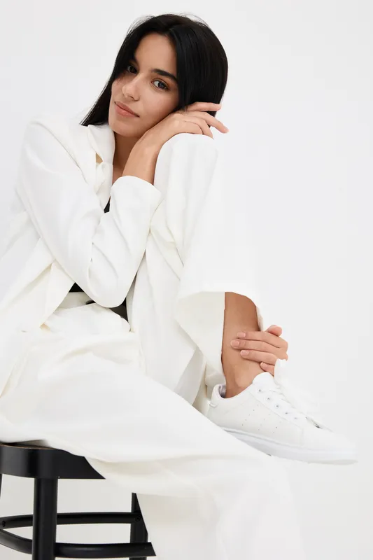 young woman in white suit and sneakers sitting on black stool, relaxed pose with natural dark hair and minimal makeup against plain white background